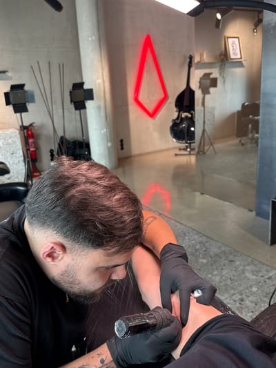 The image shows a tattoo artist working on a customer in a studio. The artist, wearing black gloves and a black shirt, is focused on the tattooing process. The customer lies on a dark surface, with their face visible. The studio has a modern design with a gray floor and walls. Various objects, including a red neon light in the shape of a triangle, a double bass, lighting equipment, and framed pictures, are visible in the background. While the exact location in Viseu, Portugal, is not specified, the image clearly depicts a tattoo studio with a contemporary aesthetic.