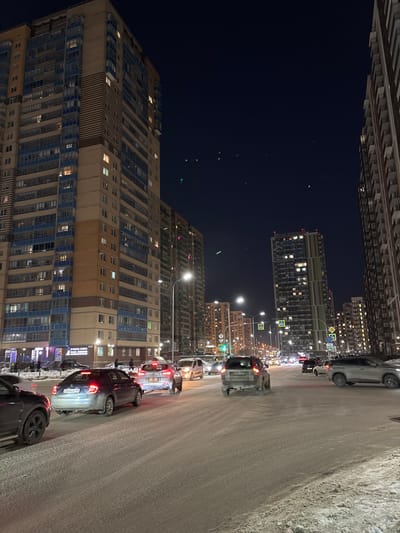 The image depicts a nighttime urban street scene in Novosergievka, Russia, under winter conditions. A multi-lane road, covered with a layer of snow or slush, occupies the foreground and mid-ground.
Several tall, multi-story residential buildings frame the scene. On the left, a prominent high-rise features light brown and blue horizontal striped facades with numerous illuminated windows. Other similar, slightly smaller high-rise buildings are visible further down the street on the left. On the right, another set of tall apartment buildings, primarily in darker tones, also display illuminated windows.
The road is populated by approximately a dozen vehicles, consisting primarily of sedans and SUVs. Most vehicles have their headlights or taillights illuminated, indicating they are either in motion or stationary in traffic. In the left foreground, a dark-colored SUV is partially visible. Immediately behind it, a silver sedan with illuminated taillights bears a partially visible Russian license plate starting with "946AB 7". To its right, another silver SUV with illuminated taillights is present. Traffic appears to be moderate, with vehicles spaced across multiple lanes.
Streetlights are brightly illuminated along the road, casting light onto the snowy surface. In the distance, traffic lights are visible, with one indicating a green signal. Standard road signs, including pedestrian crossing markers, are also discernible. The sky above is a dark, clear blue, with several small, green, elongated light artifacts scattered across it. No discernible individuals are clearly visible on the street or sidewalks.