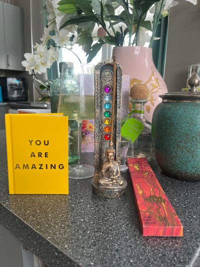 A still-life arrangement of objects is placed on a gray speckled countertop. Dominating the scene is a horizontal silver incense holder with a row of seven colorful gems and a Buddha figure at one end, holding a lit incense stick. Behind the incense holder, from left to right, are white flowers in a clear vase, a pink vase, a small clear Patron Tequila bottle, and a teal lidded jar. Adjacent to the incense holder is a yellow book with "YOU ARE AMAZING" printed on its cover in black. A red incense box with an intricate gold tree design is placed near the jar. The scene is likely set in a domestic environment, possibly in the city of Liverpool, United Kingdom, although this can't be conclusively confirmed from the image alone.