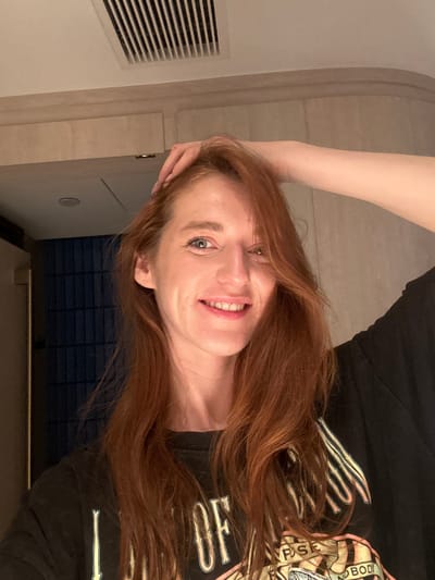 A woman with red hair smiles, looking at the camera. She wears a black t-shirt with graphic design, appearing to be in an indoor setting with a wall and ventilation grates visible. Her left hand rests on her head. The photograph is rotated at an odd angle.
