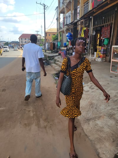 Two people are standing on a dirt path next to a road lined with shops. A young woman in a yellow and black patterned dress with pink rollers in her hair is posing with her arms outstretched. She is carrying a dark patterned bag on her shoulder. Behind her, a man in a white t-shirt and light blue jeans is facing away from the camera, holding a phone in his left hand. The scene appears to be a marketplace or a busy street in a town or city. There are shops with various goods displayed, including packaged drinks and bags. Overhead power lines are visible in the background, along with buildings and parked vehicles on the road. The lighting suggests it is daytime, with some shadows indicating sunlight. The overall atmosphere seems casual, possibly a moment captured during a walk or while shopping.