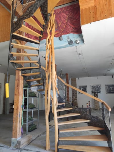 This image depicts an interior view of a spiral staircase constructed with wooden steps and a metal frame adorned with a decorative pattern. Natural, raw wooden branches are incorporated into the central support of the staircase, creating a unique organic aesthetic. The background reveals a mural on the wall featuring red curtains and a painted scene with a cat. Various artworks, including framed prints and sculptures, are visible in the surrounding space. The environment appears to be a gallery or studio, with industrial-style shelving units holding green sculptural objects. The flooring is concrete, and exposed wooden beams are present in the ceiling and walls. The lighting suggests an indoor setting with artificial lights and natural light filtering in. This scene is located at the "Kunsthofpassage" in Dresden, Germany.