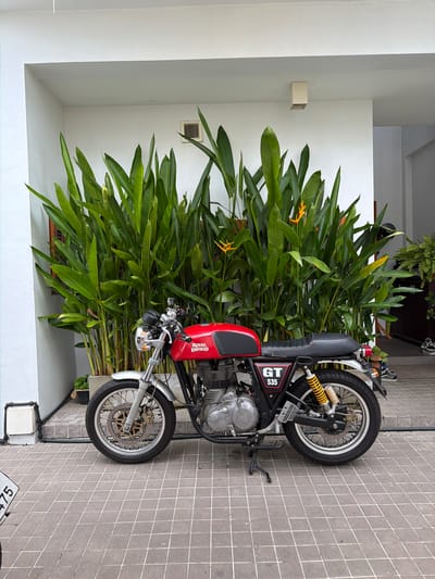 A red and silver Royal Enfield GT 535 motorcycle is parked on a tiled patio in front of a building with large green plants. The motorcycle is facing right, with its kickstand down. The building wall behind the plants is white. To the right of the motorcycle, there is a doorway leading into a darker interior, with some ferns and shoes visible. The patio is paved with square tiles. The scene is outdoors in what appears to be a residential or commercial area. The location is Chang Phueak, Thailand.