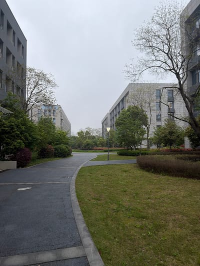This outdoor shot depicts a paved pathway curving between grassy areas and buildings in Jiangning District, China. The sky is overcast. The path is bordered by greenery, including shrubs and trees, with buildings visible on either side. The buildings have multiple windows and a modern architectural style. A street lamp is visible in the middle ground. No people are present in the scene.