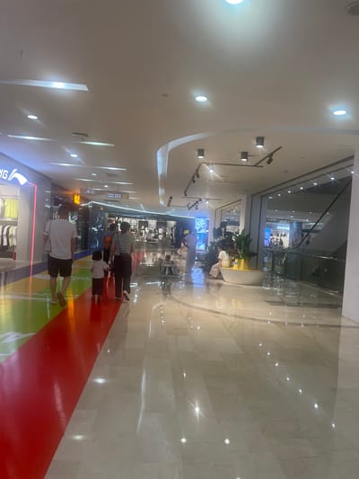 The image shows a bright, modern shopping mall interior, likely located in Liandu District, China. The floor is a polished marble, reflecting the overhead lighting and the colorful floor art. In the foreground, a man and a child are walking away from the camera. The child is a young girl wearing a light-colored t-shirt and dark pants, holding the hand of an adult. Behind them, another adult, a woman, walks with a striped shirt and dark pants. Further into the mall, other shoppers are visible, some sitting on a bench, and others browsing in stores. The stores themselves appear to be clothing retailers, with displays of apparel visible. An escalator can be seen in the background, suggesting multiple levels to the mall. The overall atmosphere is one of commerce and pedestrian activity.