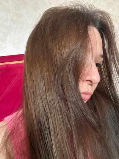 The image shows a close-up, sideways view of a young woman's face, largely obscured by her long, brown hair. She appears to be lying down, possibly on a bed or couch, indicated by the blurred pink fabric in the foreground. The woman's eye, nose, and lips are visible, and she has fair skin with some freckles. Her expression is neutral, and the lighting suggests an indoor setting, possibly during the day given the natural light suggested by the tones. The background is a textured, light-colored surface, which could be a wall or furniture. There is no visible text in the image. The overall mood is intimate and serene, focusing on the texture of the hair and the delicate features of the face.