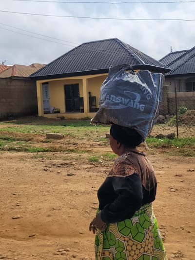 In the city of Jos, Nigeria, a woman walks with a large, dark gray bag of goods balanced on her head. She is wearing a black headwrap, a brown and black patterned top, and a green and white patterned skirt. The background shows a yellow building with a dark roof, a brick wall, and a chain-link fence. The ground is a mixture of dirt and sparse vegetation. The sky is partly cloudy.