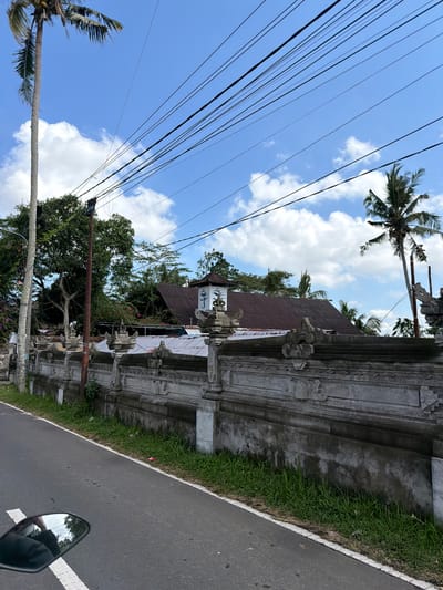 A medium shot captures a street scene in Tampaksiring, Indonesia, under a bright blue sky dotted with white clouds. A tall palm tree stands on the left, its fronds swaying gently. Utility wires crisscross the sky, supported by poles. A grey, ornate stone wall runs along the right side of the road, adorned with intricate carvings. Beyond the wall, the reddish-brown tiled roof of a building is visible, with trees and vegetation surrounding it. The road surface is asphalt, with a white line marking the edge. A motorcycle's rearview mirror is partially visible in the lower left corner, reflecting the sky and trees. The scene appears calm and quiet, suggesting an ordinary day in a tropical locale.