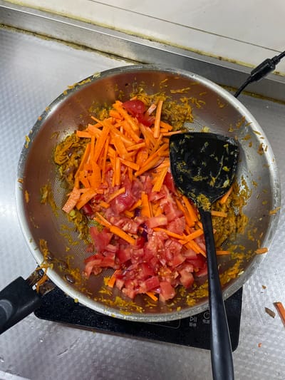 An overhead forensic view captures a cooking scene set in what is presumed to be a kitchen within Daxing District, China. The central subject is a stainless steel frying pan, approximately 25-30 cm in diameter, resting on a black induction cooktop.

The pan contains a base layer of sautéed, finely textured, yellowish-orange vegetable mixture, possibly onions or grated carrots with spices. On top of this base, two distinct piles of raw, freshly chopped vegetables are visible: a larger quantity of julienned orange carrots and a pile of diced red tomatoes. A black, flat-bladed spatula, lightly coated with the yellowish-orange mixture, is positioned within the pan, partially submerged among the vegetables.

The black induction cooktop beneath the pan displays white illuminated controls, including circular icons and Chinese characters for "暂停" (pause) and "电源" (power supply). The cooktop is situated on a light-colored, reflective countertop, possibly stainless steel or a light-colored tile, which exhibits some minor moisture or sheen.

The background consists of a light-colored wall or backsplash, also appearing to be stainless steel or tile. A horizontal seam or crevice where the wall meets the countertop shows accumulation of yellowish-brown residue. A black electrical cable is visible extending from the upper right quadrant of the image, presumably connected to the cooktop or another appliance. A small, orange piece of chopped carrot or vegetable debris is visible on the countertop to the lower right of the cooktop. No individuals are present in the frame.