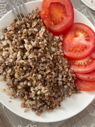 A close-up, overhead shot shows a white plate filled with cooked buckwheat and sliced tomatoes. A silver fork is partially visible, inserted into the buckwheat from the top left. The buckwheat is a mix of brown and tan grains, cooked until tender. Several bright red tomato slices are arranged on the right side of the plate, their juicy interiors exposed. The plate sits on a surface that appears to be a table with a patterned placemat. The lighting suggests it is daytime. The image was captured in Brovary, Ukraine.