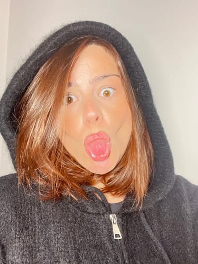 The image features a close-up of a person, likely a young adult, captured in a playful pose. The image is oriented sideways, with the person's head appearing to be tilted significantly to the left from a standard upright view. The subject is looking directly at the camera with wide, alert eyes, which are a light brown or amber color. Their eyebrows are well-defined. A key feature of their expression is their tongue sticking out slightly, adding to the humorous and lighthearted mood.

The person has shoulder-length, straight or slightly wavy hair that is a warm brown color with reddish highlights. It falls around their face and shoulders. They are wearing a dark, possibly charcoal grey or black, hooded sweatshirt or jacket. The fabric appears to be textured, possibly a fleece or knit material, and a silver zipper is visible on the front of the garment, partially open. The hood is pulled up, framing their face.

The background is uniformly plain and light-colored, suggesting a simple indoor setting, possibly a wall, which helps to keep the focus on the subject. The lighting appears bright and even, illuminating the person's features clearly. There is no visible text in the image.