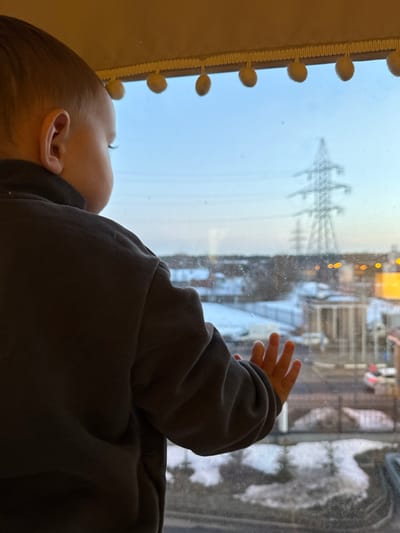 A young child, observed from behind and the right side, looks out a window in Akulovo (Gp Kubinka), Russia. The child has light brown hair and wears a dark long-sleeved garment. Their right hand is pressed flat against the windowpane. Above, a light-colored valance with spherical pom-poms decorates the window frame. The exterior scene, viewed through the window, depicts an urban or semi-urban environment with patches of snow on the ground. A street, buildings, and vehicles are visible. Multiple large metallic electricity pylons stand prominently in the distance against a pale blue sky, suggesting either early morning or late afternoon light. Distant artificial lights are visible.