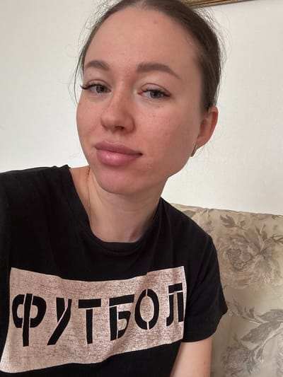 A woman is shown in a close-up, angled selfie. She has fair skin, light brown hair pulled back, and appears to be wearing minimal makeup. Her t-shirt is black with a light-colored inscription in Russian that translates to "football" or "soccer". The background consists of a plain light-colored wall on the left and a textured floral pattern on what seems to be a sofa or upholstered headboard on the right. There is no clear evidence to place her in Ферма, Russia.