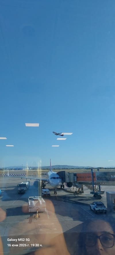 An airplane is seen taking off into a clear blue sky, its wings visible against the vast expanse. Below, on the tarmac of Valencia Airport in Manises, Spain, a large passenger jet is connected to a jet bridge. Service vehicles, including a fuel truck and a tug, are positioned near the aircraft. A car is visible further down the tarmac, and in the foreground, the reflection of a person wearing glasses can be seen on the window, superimposed over the airport scene. The date and time stamp "Galaxy M52 5G 16 юни 2026 г. 19:30 ч." is present in the lower-left corner.