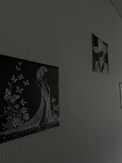 The image displays a textured wall with three artworks featuring intricate designs made of small, reflective beads. Two smaller artworks are visible in the upper left and upper center of the frame. The artwork in the upper center depicts a stylized butterfly. The larger artwork in the lower foreground is a black canvas with a design of a person and what appears to be a tree adorned with butterflies and flowers, all rendered in beads. The lighting suggests an indoor setting, possibly a home or gallery, with the light source coming from the upper left, casting subtle shadows. The beads catch the light, giving the artwork a sparkly and dimensional appearance. The overall mood is serene and artistic. No visible text is present in the image. The location context provided is Roskilde, Denmark.