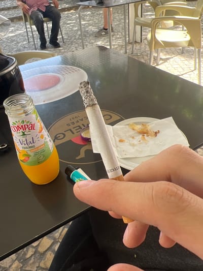 The image depicts a close-up view of a person's hand holding a lit cigarette. The scene takes place at an outdoor cafe in Almada, Portugal, as indicated by the "CAFES MELO 1937 PORTUGUES" text visible on the table. On the table, there is a bottle of Compal Vital Equilibrium Mango Laranja juice, a small lighter, and a white paper napkin with crumbs from a pastry. In the background, out of focus, a person is seated on a chair at another table, wearing a pink checkered shirt and dark pants. Several empty yellow chairs are also visible around the cafe setting. The ground is paved with cobblestones, and sunlight casts shadows across the scene.