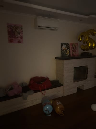 The image depicts an indoor scene with a focus on a wall decorated with artwork and balloons. On the left wall, a painting of a tiger's head surrounded by strawberries is displayed, with an air conditioning unit mounted above it. To the right of the tiger painting, two framed pictures featuring cartoon characters, one resembling Donald Duck and another a bear, are placed on a mantelpiece. A large, gold "3" shaped balloon floats above the mantelpiece. The mantelpiece itself is constructed of white stone blocks. Below the mantelpiece, on a low shelf, are several bags and clothing items, including a red duffel bag. On the floor in front of the mantelpiece are two propane tanks, one blue and one yellow, and a white balloon. The room appears to be dimly lit. The scene suggests a domestic setting, possibly decorated for a celebration.
