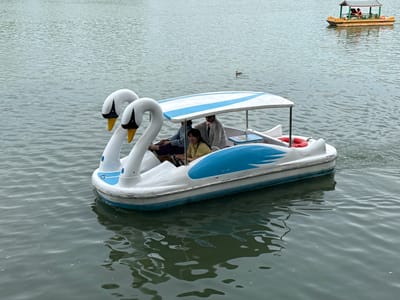 In Xicheng District, China, a white swan-shaped pedal boat navigates a body of water. Three individuals are seated within the boat; two are positioned towards the rear, while one is seated at the front. The boat features a white canopy with blue stripes, and a small blue and white table is visible. A life preserver ring is attached to the side of the boat. In the background, another boat with a striped canopy carries two individuals, and a duck swims in the water. The water's surface is rippled, reflecting the overcast sky.