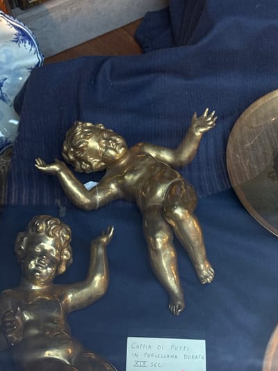 The image captures two golden-colored putti figures, likely antique or decorative items, displayed on a dark blue fabric. One putto, positioned prominently in the center, lies on its back with arms and legs extended, showcasing its chubby, infant-like form and curly hair. A small white tag is tied to its right arm. The second putto is partially visible in the bottom right, showing its head and upper body.
The dark blue fabric, possibly velvet or a similar material, serves as a backdrop, creating a contrast with the shiny gold surface of the figures. In the upper left corner, part of a circular, reflective metallic object, possibly a brass or bronze dish, is visible. Another decorative piece, a white and blue patterned ceramic bowl or plate, can be seen in the bottom left, partially covered by the blue fabric.
A white rectangular label with vertically oriented text is affixed to the display. The visible Italian inscription reads: "COPPIA DI PUTTI" (Pair of Putti), "IN PORCELLANA DORATA" (in Gilded Porcelain), and "XIX SEC." (19th Century), indicating that the figures are a pair of gilded porcelain putti from the 19th century. The setting suggests an antique shop, market stall, or display case, consistent with the location context of Como, Italy.