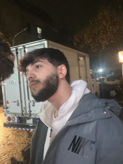 A person with dark hair and a beard is facing the camera, wearing a white hoodie and a gray Nike jacket. Behind them is a white delivery truck with a KIA logo. The license plate is partially visible, showing "8404." The setting appears to be outdoors at night, with streetlights illuminating the scene and foliage visible in the background. The ground seems wet, suggesting recent rain.