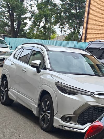 The image shows a white Toyota Corolla Cross parked on a paved street in Ussurijsk, Russia. The car is facing right, with its front end visible. A light blue fence is behind the car, and beyond that, there are trees and a brick building. A black SUV is partially visible to the right of the Corolla Cross, and a red taillight of another vehicle is in the foreground. The license plate on a white vehicle to the left of the Corolla Cross is partially visible and reads "BK 12S". The scene appears to be an outdoor parking area or street.