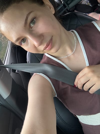 A woman is seated in a car, wearing a brown and white top. She has a necklace on and her seatbelt is fastened across her chest. Her hand is visible, resting on the seatbelt. The image is taken from a slightly elevated angle, looking down at her face. Her eyes are open and she appears to be looking towards the camera. The interior of the car is visible, including the black leather seats and the grey seatbelt. The background is blurred, but appears to be an outdoor setting with greenery. The lighting suggests it is daytime. There is no visible text in the image.