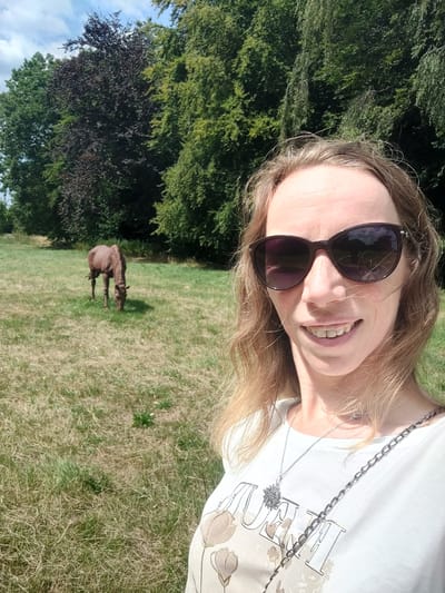 A blonde woman with sunglasses is taking a selfie in a grassy field. A brown horse is grazing in the background. The scene is set in a wooded area with green trees and a blue sky with clouds. The location is the city of Isselburg, Germany.