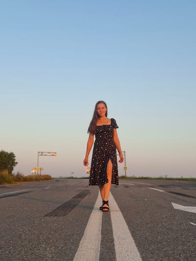 A young woman with long brown hair walks towards the camera on a paved road. She is wearing a black floral-print dress with a thigh-high slit and black sandals. The road has white lane markings, and there are tire tracks visible on the asphalt. In the background, there are signs and vegetation. The sky is clear and a pale blue, suggesting it is either sunrise or sunset. The location is identified as Perevoznoe, Russia.
