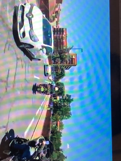 The image displays a daytime urban street scene in Kafr Sousa Municipality, Syria, captured from the perspective of a vehicle, likely a motorcycle. In the foreground, the upper body of a person wearing dark attire and a light-colored helmet is visible, positioned on a dark-colored motorcycle with a visible windshield. On the asphalt road ahead, a dark-colored motorcycle with a substantial front fairing and a rear storage compartment is centrally located. Further ahead and to the right, a white four-door sedan with tinted rear windows is present; its license plate is partially visible, commencing with "85". A smaller white vehicle is positioned behind the dark motorcycle and to the left of the white sedan. The road features visible white lane markings. Along the right side of the road, multiple multistory buildings constructed from brown or reddish-brown brick are visible, interspersed with numerous mature green trees and a dark-colored street light pole. The upper portion of the scene is dominated by a clear, bright blue sky.