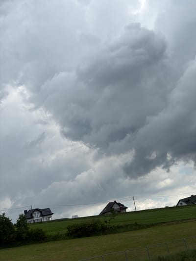The image depicts a cloudy, overcast sky dominating the upper portion of the frame. Below the sky, a green, hilly landscape stretches across the middle and lower sections. Three houses are visible on the crest of the hill. The house on the left is a modern, multi-story building with a dark roof and white walls, featuring several windows and balconies. A single utility pole with wires runs in front of it. To the right of this house, another house is partially visible, also with a dark roof and white structure. Further to the right, the peak of a third house with a green roof is visible. The foreground is a grassy field, with a portion of a metal fence at the bottom. The scene appears to be a rural or semi-rural residential area, possibly in the vicinity of Męcina, Poland, given the style of the houses and the landscape. There are no people visible in the image.