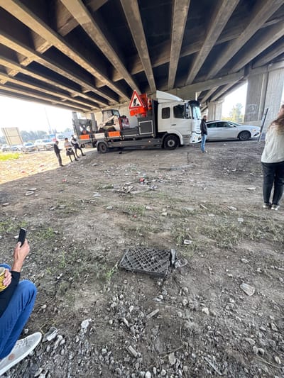 The scene depicts an unpaved area beneath a concrete overpass in Rosh HaAyin, Israel, under daylight conditions. The ground consists of dry, uneven dirt mixed with gravel, sparse green vegetation, and significant scattered litter including plastic fragments and cardboard.

A white Mercedes-Benz flatbed truck, license plate number 979-331 (Israeli plates), is parked centrally on the dirt. Its flatbed carries a white compact construction machine, possibly a mini excavator or skid steer loader. An orange triangular warning sign with a black worker silhouette and a red "500" box is affixed to the truck's load. To the right of the truck, a white sedan is parked facing the same direction. In the background, on a paved surface, a line of additional vehicles is visible.

Several individuals are present. On the left side, a group of three to four men stand conversing near the front of the truck's flatbed. Another man in a dark jacket and jeans stands alone near the passenger side of the truck. On the far right, partially visible from the rear, is a woman with long hair wearing a light-colored top and dark pants. In the immediate foreground, to the bottom left, a person's lower body is visible; they are holding a black smartphone with the screen facing away and a yellow-striped cup. A black, grid-patterned plastic or rubber grate-like object lies on the ground in the mid-foreground.

The overhead structure consists of numerous parallel concrete beams forming the underside of the multi-lane overpass.