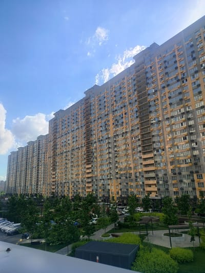 A low-angle, wide shot captures a long, modern apartment building with a muted orange and gray facade under a bright blue sky with scattered white clouds. The building extends across the frame, with numerous windows and balconies visible. In the foreground, a lush green area with trees and neatly trimmed bushes is situated in front of the building. A parking lot with several cars and a motorcycle is visible to the left of the green space. The overall scene depicts a residential area with contemporary architecture and landscaping. The location of Labinsk, Russia, is not visually discernible from the image.