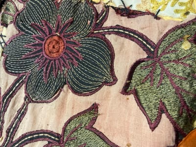 The image shows a close-up of an embroidered textile. The embroidery depicts a floral design with large, dark petals outlined in purple and white stitching, and a reddish-brown center. There are also green embroidered leaves with purple outlines. The background fabric is a pale pink, and some areas show signs of age and wear, with small stains and loose threads. The overall impression is of a detailed, vintage piece of embroidery. This image does not depict any location in Sabadell, Spain, as it focuses solely on the textile itself.