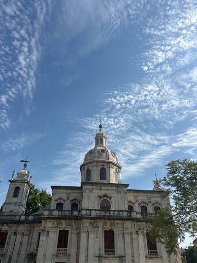This media file features a view of the Estrela Basilica in Lisbon, Portugal, on a clear day. The camera angle is tilted upwards, capturing the bright blue sky filled with wispy white clouds. The basilica's grand dome and a bell tower are prominent elements, rendered in light-colored stone with architectural details like arched windows and decorative carvings. Some green trees are visible, adding a touch of nature to the scene. The sunlight suggests it is daytime. The overall mood is serene and awe-inspiring, highlighting the architectural grandeur of the basilica against the expansive sky. No people are visible in the frame, and there is no visible text.