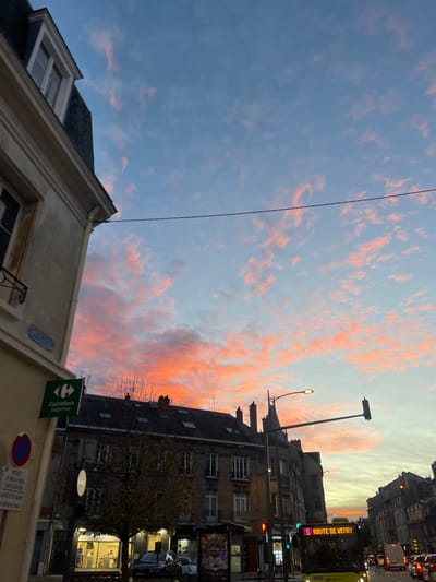 The image depicts an urban street scene in Reims, France, at either dusk or dawn, characterized by a vibrant sky. The upper portion of the frame is dominated by a clear blue sky transitioning to light blue and then a brilliant orange, pink, and red at the horizon, with scattered, fiery-hued altocumulus clouds.
In the immediate foreground on the left, a light-colored building with a classic Haussmannian-style mansard roof and dormer windows is visible. A green "Carrefour express" sign is mounted on its lower facade, along with a partially obscured blue street sign and a "no parking" road sign.
The center and right portions of the image showcase a street lined with multi-story buildings, predominantly dark grey, featuring multiple windows and pitched roofs with chimneys. Bare deciduous trees are situated along the street. A bright yellow public transit bus, displaying "L5 ROUTE DE VITRY" on its front destination sign, moves rightward with its headlights illuminated. Other vehicles, including cars and a white van, follow in the distance, their rear lights glowing red. Streetlights, one actively emitting light, and traffic lights (showing both red and green signals) are positioned along the thoroughfare. Overhead power lines or tram lines traverse the sky horizontally. A bus stop shelter with an advertisement is visible on the sidewalk.