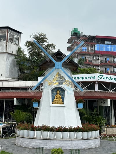The image depicts a windmill structure with blue blades and a Buddha statue on its side, labeled "Annapurna Garden," likely a decorative element or landmark within a garden or park. The windmill is white with painted details. The statue has golden hues. The surrounding environment consists of buildings with varying architectural styles, including structures with red roofs, air conditioning units, and glass windows. The sky is overcast. The scene is located in the city of Pokhara, Nepal. The details provide an objective view of the image's contents and its implied location.