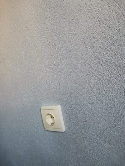 The image displays a light blue-grey wall with a uniform, finely textured surface, characterized by irregular, short, undulating ridges. Positioned in the lower-middle left quadrant of the wall is a single white electrical wall socket. The socket, designed for European Type F (Schuko) plugs, features two circular pinholes and upper/lower grounding clips. It is slightly tilted clockwise relative to the vertical axis of the image frame. No plug is inserted into the socket, and its internal components are visible. The wall surface exhibits slight variations in brightness, appearing marginally lighter in the upper left section.