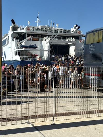 This media file depicts a group of people disembarking a ferry or ship. The scene is set in a sunny, outdoor environment, indicated by the bright blue sky and clear shadows cast by the metal grate in the foreground. The time of day appears to be daytime, likely late morning or afternoon.

The subjects are a large number of people, seen from the chest up as they stand behind a metal grate. They are dressed in casual clothing, suggesting a travel or leisure context. Many are looking towards the camera or in the direction they are moving. Some appear to be holding phones, possibly taking pictures or videos. The overall mood seems neutral to slightly excited, typical of travelers arriving at a destination.

In the background, the large white structure of a ferry or ship is visible, with multiple decks, railings, and what appear to be smokestacks or ventilation shafts. A bus is partially visible at the top of the frame, with branding that includes "NST Travel," "Santorini," and "Athens," along with phone numbers. There is also a license plate visible on a vehicle in the background with "EMT 88" on it. The presence of the ship, bus, and travel agency information strongly suggests a port or ferry terminal.

Visible text includes "CHAMPIONS LEAGUE JET," "NST Travel," "Santorini-30 22860 71199," "Athens)+30 210 3390660," and "www.nsttravel.com." The location context is further reinforced by the mention of Santorini and Athens. The scene is likely depicting a busy arrival or departure point for passengers traveling between Greek islands or the mainland.