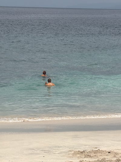 Two individuals are swimming in the ocean near Manggis, Indonesia. The person closer to the foreground is a man with his back to the camera, his head and shoulders visible above the water. He is swimming in clear, turquoise water. Further out in the deeper, grayish-blue ocean, a second individual, appearing to be a woman with short hair, is also swimming. The shoreline, with its light-colored sand and gentle waves, is visible in the bottom portion of the image. The sky is overcast, creating a muted light.