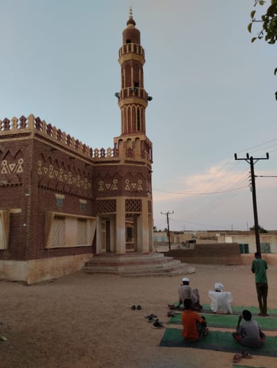 A brick mosque with a tall minaret dominates the scene under a pale sky. The mosque features geometric patterns along its edges. Several people are praying on mats outside the mosque. Additional shoes are scattered on the ground nearby. In the background, there are simple village buildings and utility poles with wires. The ground appears to be sandy or dirt.