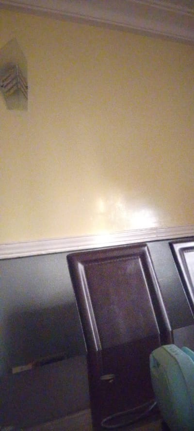 The image shows an interior scene, possibly a room. The wall is painted a pale yellow color and has a white crown molding at the top. Below the molding is a dark gray surface, which could be a wall, a cabinet, or a piece of furniture.

In the foreground, there is a dark brown chair with a carved panel. To the right of the chair, a portion of another chair or furniture piece is visible, with a lighter color. On the far right, a light blue object, possibly a cushion or a bag, is partially visible.

On the far left of the image, near the top of the pale yellow wall, there is a light-colored object mounted on the wall. It appears to be a square or rectangular fixture with angled slats or grooves.

The image is somewhat grainy and appears to be taken in low light conditions. There are no people visible in the frame. The overall impression is that of a sparsely furnished room. The location in Madalla, Nigeria, is not explicitly depicted by any landmarks or context within the image itself.