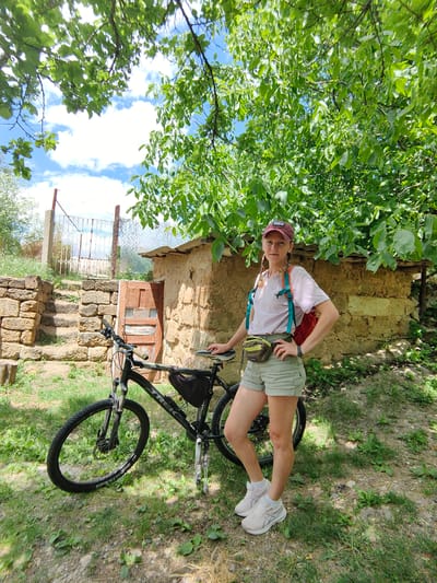 A young woman with blonde hair braided into two pigtails stands next to a black Trek mountain bike. She is wearing a maroon baseball cap, a light pink t-shirt, light green shorts, and white athletic shoes. A small brown fanny pack is around her waist, and a red backpack is slung over her right shoulder. The bike has a black saddlebag attached to its frame.
The woman is positioned in front of a rustic, earthy-toned building with a weathered wooden door. To the left of the building, a stone staircase leads up to a metal gate. The scene is outdoors, with lush green trees and foliage overhead, suggesting a rural or natural setting. The ground is covered in grass and dirt. The sky, visible through the tree branches, is partly cloudy and blue. The overall impression is a sunny day in a somewhat rustic, possibly Ukrainian, rural environment within the Zuyivka rural settlement.