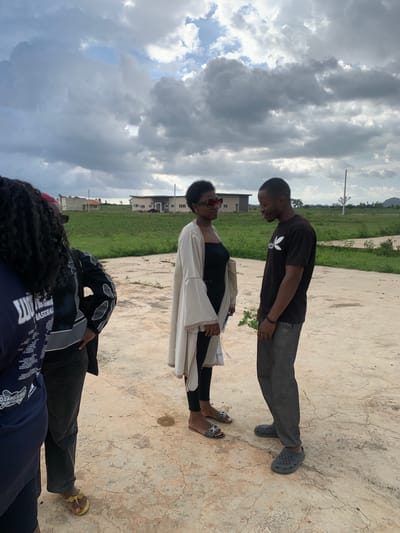 The media file depicts a group of individuals outdoors in Gwafan, Nigeria. The setting appears to be an open, paved area with grassy fields and some buildings in the background. The sky is filled with dramatic, cloudy formations, suggesting overcast or potentially stormy weather.
The subjects include at least three people. In the foreground, a person wearing a dark blue shirt with white text and intricate graphics is partially visible. Behind them, another person is seen from the waist down, wearing dark pants and black and grey patterned outerwear, with one foot in a yellow sandal. Further back, two people are more fully visible, facing each other. One, wearing a black t-shirt and grey pants with grey crocs, looks down. The other, wearing a black top and a cream-colored garment over it, with red-framed sunglasses, looks up.
The activity seems to be a casual gathering or interaction among the individuals. The image is taken in daylight, given the illumination. The cloudy sky creates a somewhat dramatic atmosphere. The architecture of the buildings in the background hints at a residential or developing area. There are no overt signs of a specific event, but the poses suggest conversation or a shared moment.
Visible text on the shirt in the foreground includes "GRAND" and other words that are difficult to discern clearly due to the angle and resolution. The overall impression is one of everyday life in an outdoor Nigerian environment under a dynamic sky.