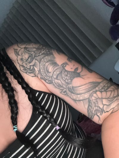 The image shows a close-up of a person's arm with a large tattoo of a woman intertwined with a snake and a flower. The person is wearing a black and white striped top. Their dark hair is braided and falls over their shoulder. The background appears to be a wall with horizontal ribbed texture, possibly acoustic paneling. The lighting is dim. The image is a photograph taken indoors. The location in the city of Kuklen, Bulgaria is not discernible from the image content.