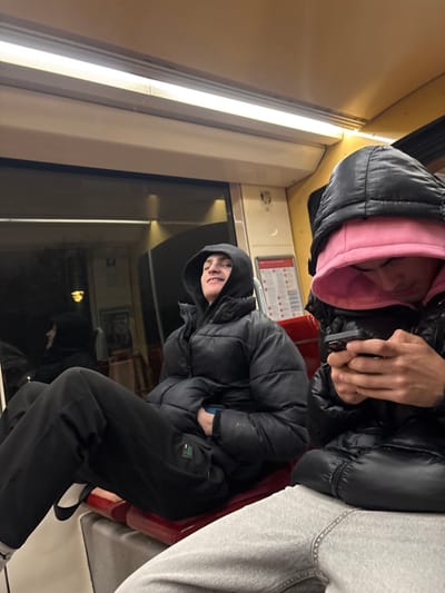 Two people are on a train. One person wears a black puffer jacket with a pink hoodie underneath. The other wears a black puffer jacket with black pants and white sneakers. They sit next to a train window. The train's interior includes red seats and lights. There is a safety information sticker.
