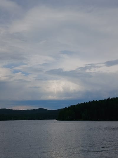The image depicts a tranquil lake scene with a dark, forested shoreline under a cloudy sky. The water is calm, with subtle ripples reflecting the muted light. Hills covered in dense green trees form the backdrop, creating a sense of depth and wilderness. In the distance, a small red boat is visible on the water, adding a touch of human presence to the otherwise natural landscape. The sky is filled with a mix of gray and white clouds, with hints of blue peeking through, suggesting an overcast or potentially approaching weather system. The overall atmosphere is peaceful and serene, evoking a sense of natural beauty and quietude. The location is identified as the city of Revda, Russia, a place known for its natural landscapes and bodies of water, which align with the scene presented.