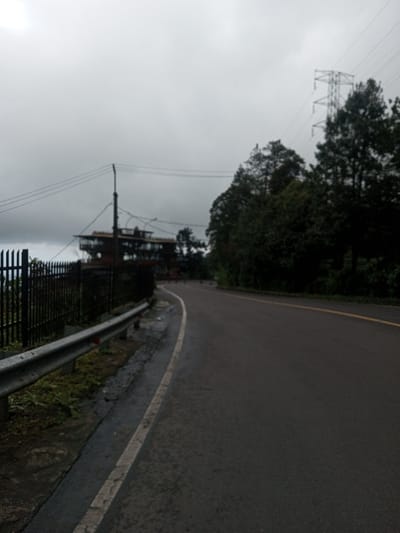 The image depicts a winding, wet asphalt road in Bangtsho, Bhutan, under an overcast grey sky. The road features a solid white line marking its left edge and a yellow line further right indicating lane separation. Along the immediate left side of the road, a silver metal guardrail with a red reflector is present, bordering a strip of damp grass and earth. Behind the guardrail, a dark metal fence with pointed vertical bars runs parallel to the road. Beyond the fence, a multi-story, dark-colored structure, possibly a building under construction or renovation, is visible on an elevated position. Utility poles with overhead wires traverse the scene, with one pole positioned near the fence line on the left. The right side of the road is flanked by a dense forest of dark green trees. In the upper right background, a tall metal lattice transmission tower is discernable above the treeline, connected by multiple overhead power lines that extend across the frame. No individuals or vehicles are visible in the scene.