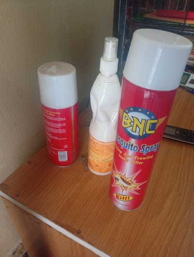 The image shows three aerosol cans and a spray bottle on a wooden surface. The aerosol cans are red and white, with one featuring the text "BNC Mosquito Spray Flying And Crawling Insect Killer." Another can has text that is partially visible and reads "And Crawling Insect Killer." The spray bottle is white and appears to be a disinfectant or cleaning product. The background is a wall and a shelf with books. The scene is indoors, suggesting a domestic or commercial setting. The objects are arranged on a wooden table or shelf. The lighting appears to be natural or artificial, casting shadows. The image does not provide any specific location information within Zanzi, Nigeria.
