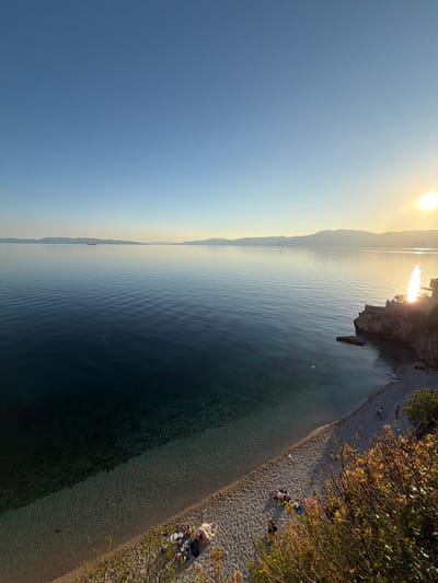 The image captures a scenic view of the Adriatic coast in Grad Rijeka, Croatia. The sun shines brightly, casting a glow across the water and sky. In the distance, the coastline stretches along the horizon. Closer to the viewer, a rocky outcrop juts into the sea. A narrow pebble beach lines the coast, and several people are relaxing or sunbathing near some trees. The water appears clear.