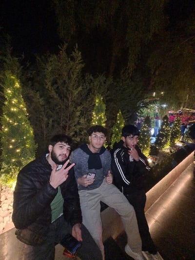 Three young men pose for a photo outdoors at night. A man with a beard closest to the camera is throwing up a peace sign. A second man behind him looks at his phone. The third man is furthest from the camera. The men are either sitting or lying on a ledge. Behind the men are trees adorned with lights. In the distance are more lights and structures that are out of focus.
